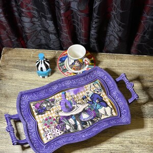 May include: Purple serving tray with ornate floral design and a whimsical Alice in Wonderland scene featuring the Mad Hatter, the White Rabbit, and Alice. The tray has two handles and is perfect for serving tea or snacks.