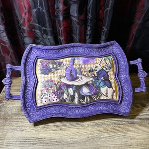 May include: A purple serving tray with an ornate frame and a whimsical Alice in Wonderland themed design. The tray features a scene from the story with Alice, the Mad Hatter, and the White Rabbit.