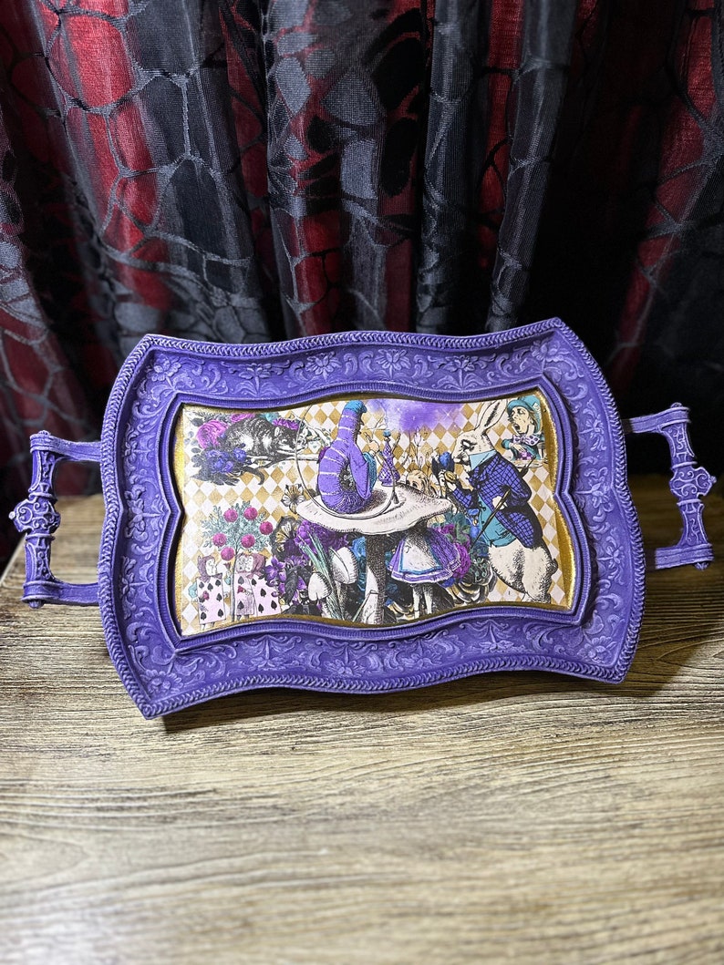 May include: A purple serving tray with an ornate frame and a whimsical Alice in Wonderland themed design. The tray features a scene from the story with Alice, the Mad Hatter, and the White Rabbit.