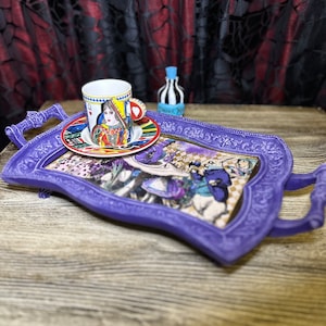 May include: A purple serving tray with a whimsical Alice in Wonderland design. The tray features a teacup with a Queen of Hearts design and a small bottle with a striped design.