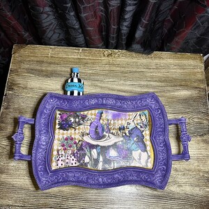 May include: Purple serving tray with ornate edges and a whimsical Alice in Wonderland design featuring the Mad Hatter, the Queen of Hearts, and the Cheshire Cat. The tray has two handles and a small bottle with the text "Drink Me" on it.