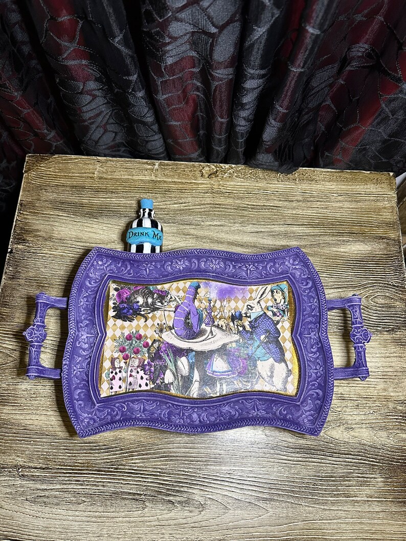 May include: Purple serving tray with ornate edges and a whimsical Alice in Wonderland design featuring the Mad Hatter, the Queen of Hearts, and the Cheshire Cat. The tray has two handles and a small bottle with the text "Drink Me" on it.