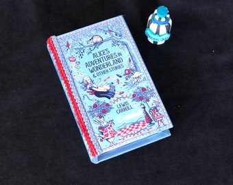 Puede incluir: Un libro azul con un borde rojo y blanco. La portada del libro presenta ilustraciones de personajes de Alicia en el País de las Maravillas y el título "Alice's Adventures in Wonderland & Other Stories" de Lewis Carroll.