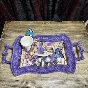 May include: Purple serving tray with a whimsical Alice in Wonderland design featuring characters like the Mad Hatter, the Queen of Hearts, and the White Rabbit. The tray has two handles and a white ceramic mug with a blue rim.