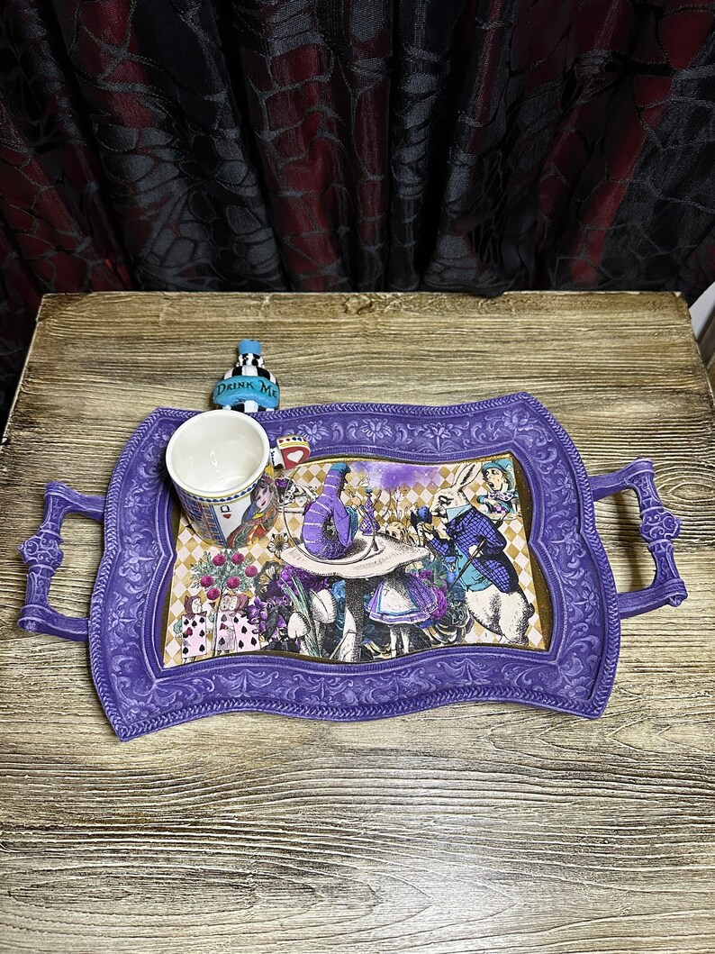 May include: Purple serving tray with a whimsical Alice in Wonderland design featuring characters like the Mad Hatter, the Queen of Hearts, and the White Rabbit. The tray has two handles and a white ceramic mug with a blue rim.