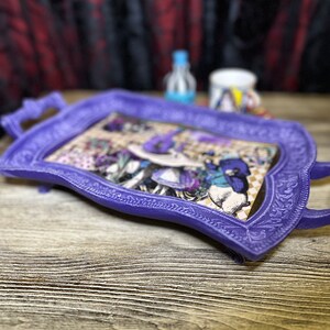 May include: A purple tray with an ornate design and a vintage illustration of a woman in a hat. The tray is on a wooden surface.