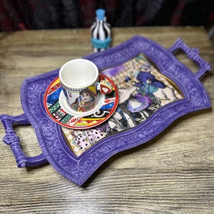May include: A purple serving tray with a decorative, ornate border and a handle on each side. The tray features a colorful Alice in Wonderland themed design with a white teacup and saucer on top.