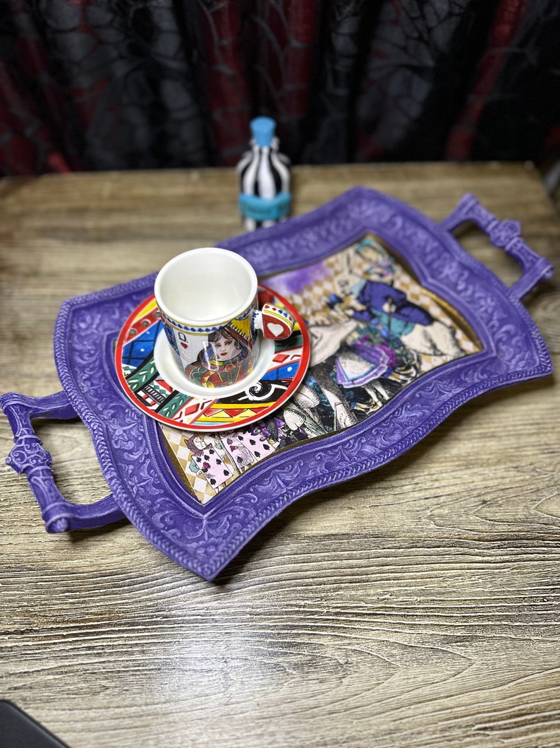 May include: A purple serving tray with a decorative, ornate border and a handle on each side. The tray features a colorful Alice in Wonderland themed design with a white teacup and saucer on top.