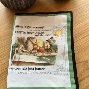 May include: A green and black fabric tea cozy with a white illustration of Alice in Wonderland characters having tea. The text reads "Two days wrong! I told you butter wouldn't suit the works. It was the best butter. The March Hare meekly replied."
