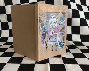 May include: A brown cardboard box featuring a colourful illustration of a girl in a white dress and a red and white house. The illustration also includes a white rabbit, mushrooms, and butterflies.