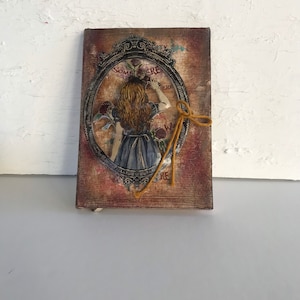 Könnte beinhalten: Eine Vintage-Rahmenillustration eines Mädchens mit langen blonden Haaren, das ein blaues Kleid trägt und eine Blume hält. Der Rahmen ist verziert und trägt die Aufschrift "Somewhere" und "Wonder". Der Hintergrund ist in einem verwitterten Braun- und Rottönen gehalten.