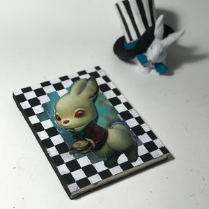 Könnte beinhalten: Ein schwarz-weiß kariertes Notizbuch mit einem weißen Kaninchen, das eine Taschenuhr auf dem Cover hält. Das Kaninchen trägt eine rote Weste und hat rote Augen.