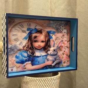 May include: Decorative rectangular tray with a blue border and handles. The tray features an illustration of a girl in a blue dress holding a rabbit, with a clock and floral background. The tray's edge has a black and white checkered pattern.
