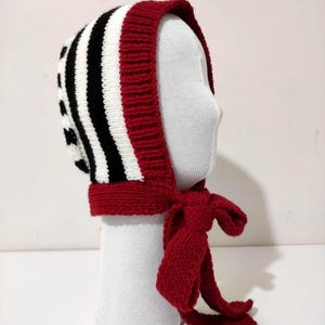 May include: A knitted bonnet with a black and white striped pattern, edged with red. The bonnet has a red tie that fastens under the chin. The item is displayed on a white mannequin head.