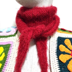 May include: A vibrant red fuzzy knit scarf tied in a simple knot, layered over a garment constructed from multi-colored crochet granny squares featuring green, yellow, orange, and white floral patterns on a cream-colored base.