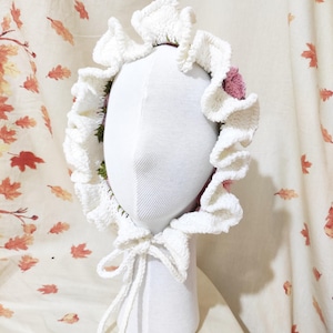 May include: A white, crocheted bonnet with a ruffled brim and a tie closure. The bonnet is adorned with pink and green floral accents. The bonnet is displayed on a white mannequin head against a backdrop of autumn leaves.
