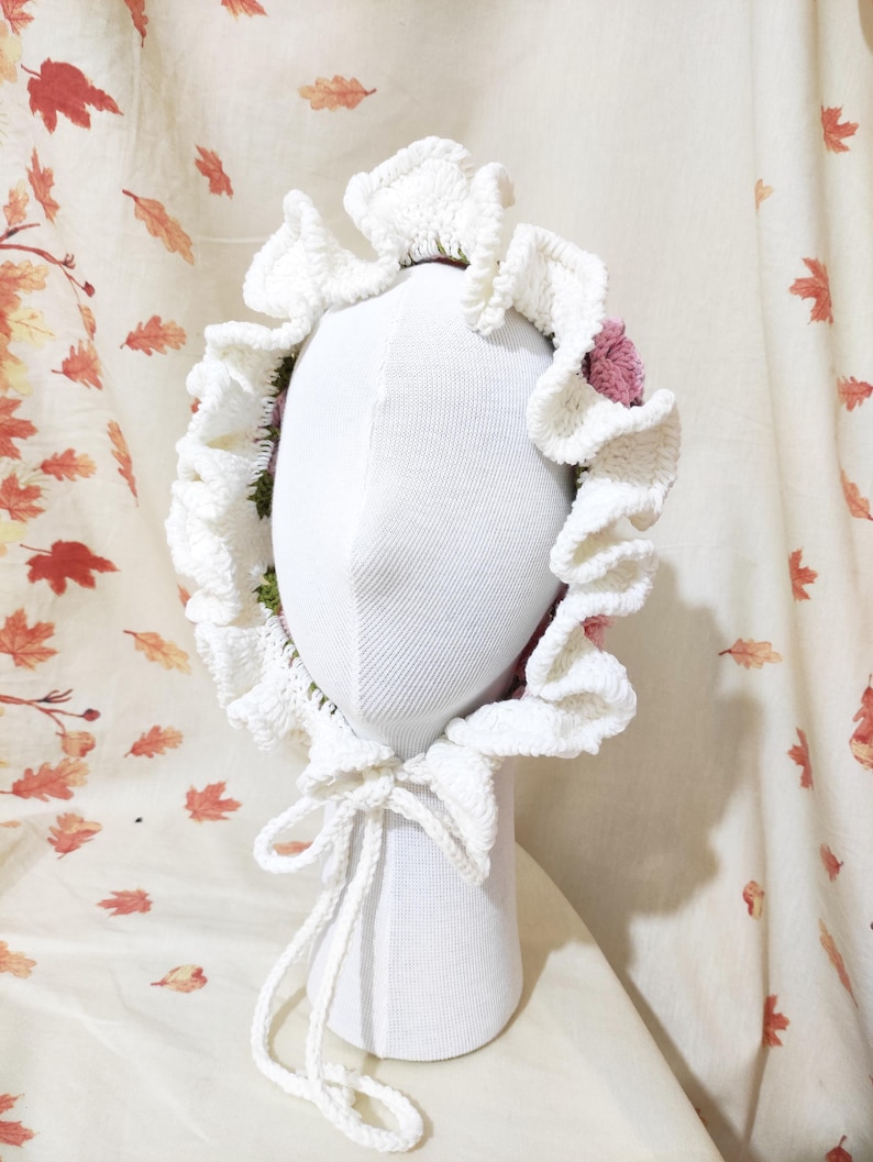 May include: A white, crocheted bonnet with a ruffled brim and a tie closure. The bonnet is adorned with pink and green floral accents. The bonnet is displayed on a white mannequin head against a backdrop of autumn leaves.