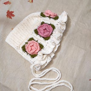 May include: A white crochet bonnet with pink and green floral accents. The bonnet features three square floral designs and a ruffled edge. White ties are attached for securing the bonnet.