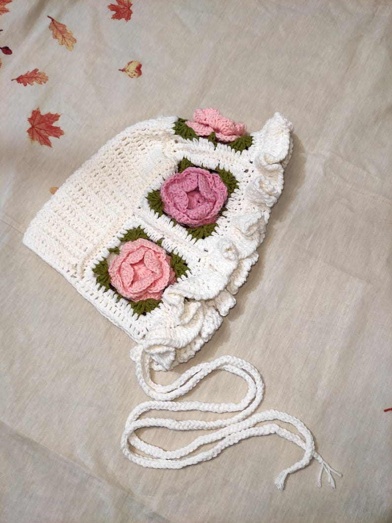 May include: A white crochet bonnet with pink and green floral accents. The bonnet features three square floral designs and a ruffled edge. White ties are attached for securing the bonnet.