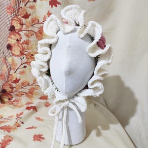 May include: A white crochet bonnet with a ruffled brim and a pink accent. The bonnet is tied with a white cord and is displayed on a white mannequin head. The background features a fall-themed fabric with leaves and pumpkins.