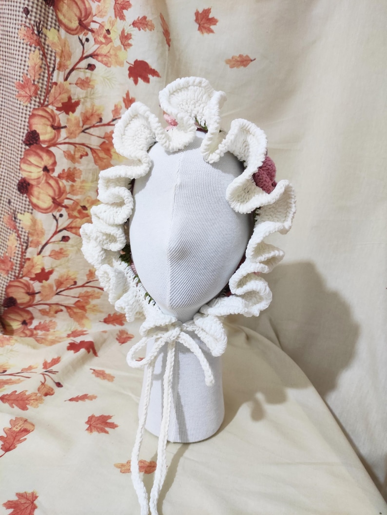 May include: A white crochet bonnet with a ruffled brim and a pink accent. The bonnet is tied with a white cord and is displayed on a white mannequin head. The background features a fall-themed fabric with leaves and pumpkins.