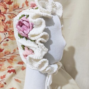 May include: A white crochet bonnet with a floral design. The bonnet features a large, ruffled brim and is adorned with pink and green crocheted flowers. The bonnet is displayed on a white mannequin head.