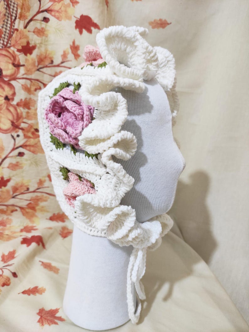 May include: A white crochet bonnet with a floral design. The bonnet features a large, ruffled brim and is adorned with pink and green crocheted flowers. The bonnet is displayed on a white mannequin head.