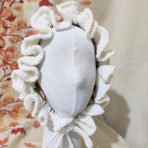 May include: A white crocheted bonnet with ruffled edges and a bow tie closure. The bonnet is displayed on a white mannequin head. The interior of the bonnet features a floral pattern with pink and green accents. The background has a fall-themed pattern.