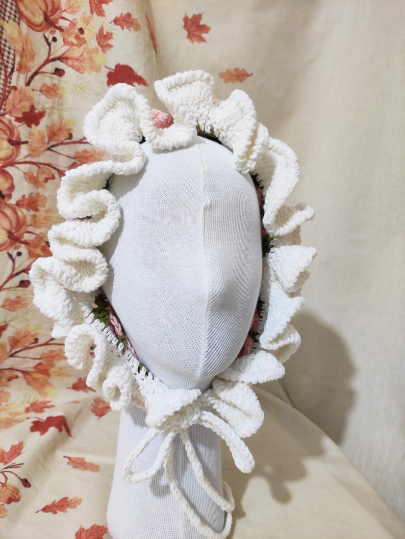 May include: A white crocheted bonnet with ruffled edges and a bow tie closure. The bonnet is displayed on a white mannequin head. The interior of the bonnet features a floral pattern with pink and green accents. The background has a fall-themed pattern.