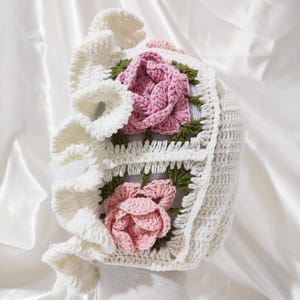 May include: A white crochet bonnet with a ruffled brim and two square floral appliqu&eacute;s. Each appliqu&eacute; features a pink rose with green leaves. The bonnet is designed with a textured stitch and a tie closure.