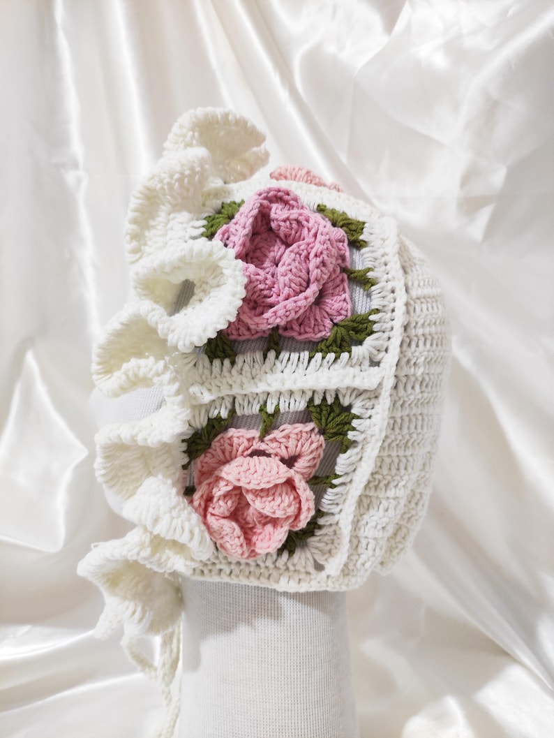 May include: A white crochet bonnet with a ruffled brim and two square floral appliqu&eacute;s. Each appliqu&eacute; features a pink rose with green leaves. The bonnet is designed with a textured stitch and a tie closure.