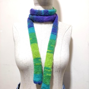 Puede incluir: Bufanda tejida a mano en tonos azules, verdes y morados. La bufanda se exhibe en un maniquí, mostrando su diseño a rayas de colores. La textura de la bufanda parece suave y cálida, perfecta para el clima frío.