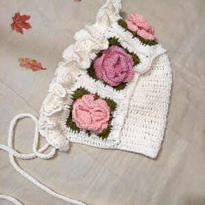 May include: A handmade white crochet bonnet with pink rose appliqu&eacute;s and green leaves. The bonnet features a ruffled edge and ties. The floral design is centered on the front and sides of the bonnet. The bonnet is made of soft yarn.