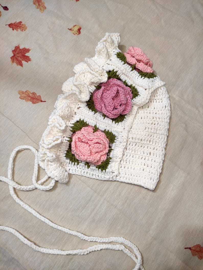 May include: A handmade white crochet bonnet with pink rose appliqu&eacute;s and green leaves. The bonnet features a ruffled edge and ties. The floral design is centered on the front and sides of the bonnet. The bonnet is made of soft yarn.
