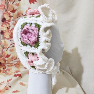 May include: A white crochet bonnet with a square floral design featuring a pink rose and green leaves. The bonnet has a ruffled brim and is displayed on a mannequin head. The background has a fall-themed fabric.