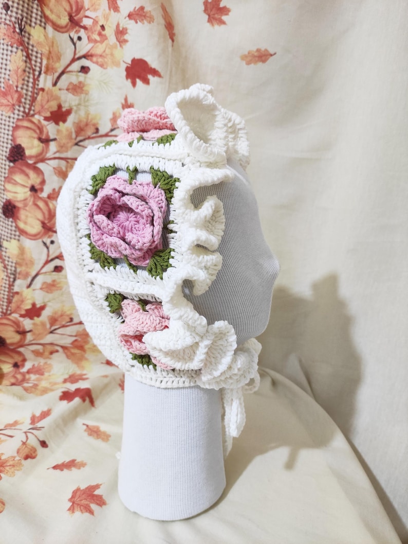 May include: A white crochet bonnet with a square floral design featuring a pink rose and green leaves. The bonnet has a ruffled brim and is displayed on a mannequin head. The background has a fall-themed fabric.