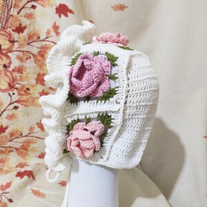 May include: A white crochet bonnet with a ruffled edge and ties. The bonnet features square panels with pink rose appliqu&eacute;s and green leaves. A small pink rose is on top. The bonnet is displayed on a white form.
