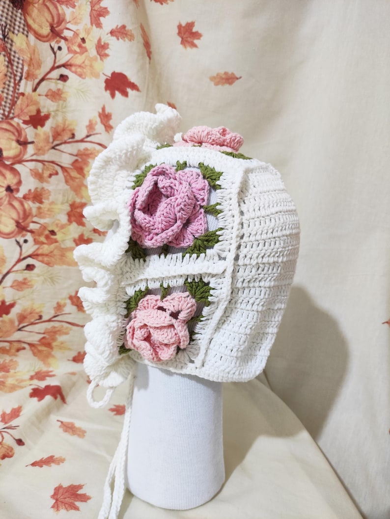 May include: A white crochet bonnet with a ruffled edge and ties. The bonnet features square panels with pink rose appliqu&eacute;s and green leaves. A small pink rose is on top. The bonnet is displayed on a white form.