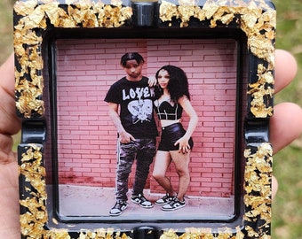 May include: Black ashtray with gold leaf accents. The ashtray has a square shape and a picture of a couple in the center. The picture shows a man and a woman standing in front of a brick wall. The man is wearing a black t-shirt with the word "Lover" printed on it. The woman is wearing a black crop top and black shorts.