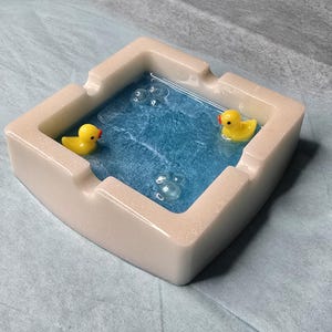 May include: A square ashtray with a light beige exterior and a blue interior resembling a pool. Two yellow rubber duckies float in the blue water, with clear bubble accents. A unique and decorative item.