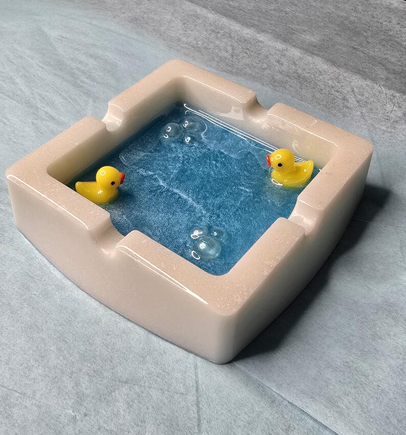 May include: A square ashtray with a light beige exterior and a blue interior resembling a pool. Two yellow rubber duckies float in the blue water, with clear bubble accents. A unique and decorative item.