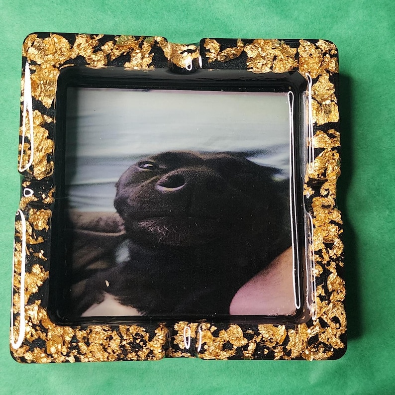 Op de afbeelding: Zwarte vierkante asbak met een bladgouddesign rond de rand. De asbak heeft een foto van een zwarte hond erin.