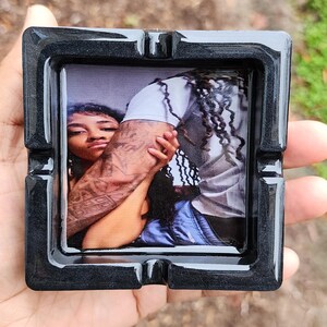 Op de afbeelding: Een zwarte vierkante asbak met een foto van een stel dat elkaar omhelst. De foto toont een vrouw met lang donker haar en een man met lange dreadlocks. De man heeft tatoeages op zijn arm.