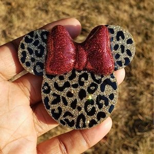 May include: A gold and black leopard print Minnie Mouse hair bow with a red glitter bow.