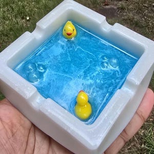 May include: A white square ashtray with a blue resin interior, featuring two yellow rubber duckies. The ashtray has a glossy finish and is held in a hand, with green grass in the background.