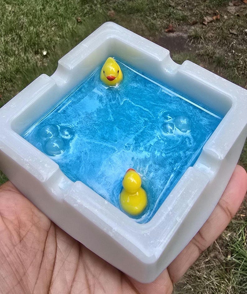 May include: A white square ashtray with a blue resin interior, featuring two yellow rubber duckies. The ashtray has a glossy finish and is held in a hand, with green grass in the background.