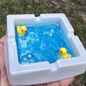 Op de afbeelding: Een witte vierkante asbak met een blauw harsinterieur, met twee gele badeendjes en bubbelaccenten. De asbak wordt in een hand gehouden, met een grasachtige achtergrond zichtbaar.