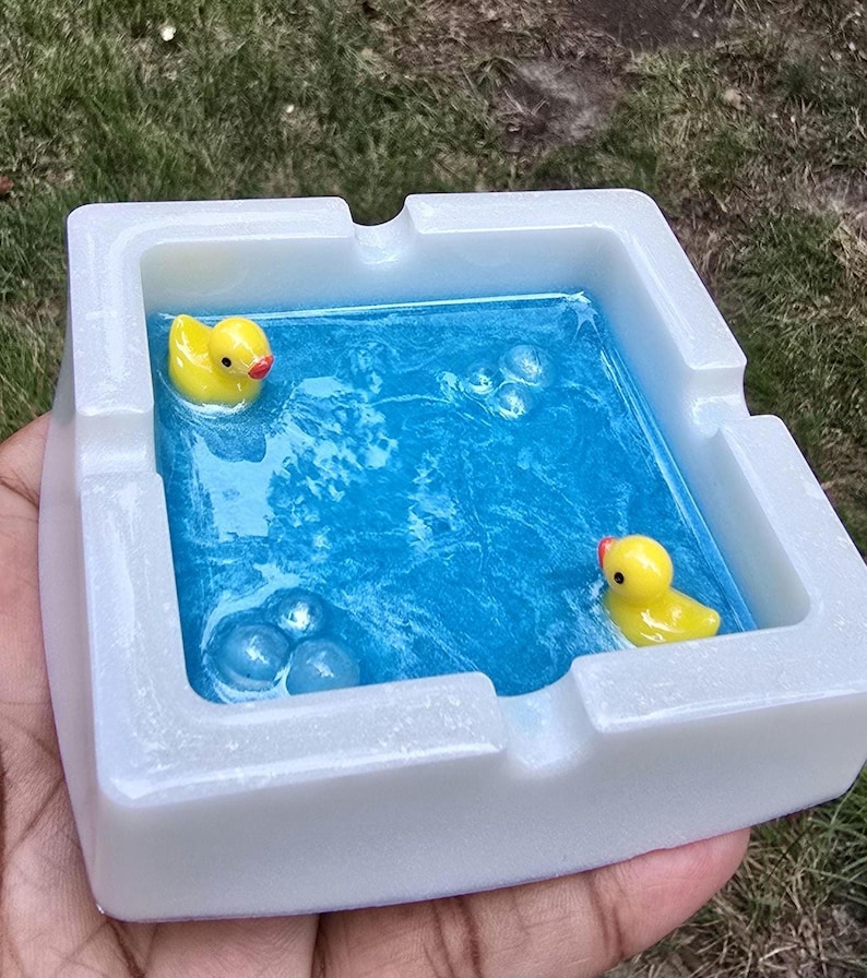 May include: A white square ashtray with a blue resin interior, featuring two yellow rubber duckies and bubble accents. The ashtray is held in a hand, with a grassy background visible.