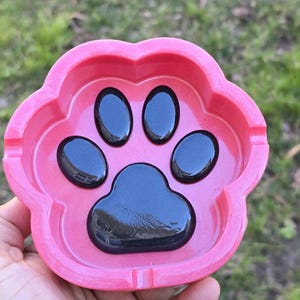 Op de afbeelding: Roze asbak in de vorm van een hondenpoot met een zwart hondenpootontwerp.