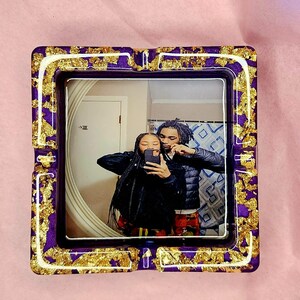 Op de afbeelding: Een paarse en gouden asbak met een foto van een stel erin. De foto toont een man en een vrouw die naar de camera lachen. De asbak heeft een witte rand en gouden vlokken overal.